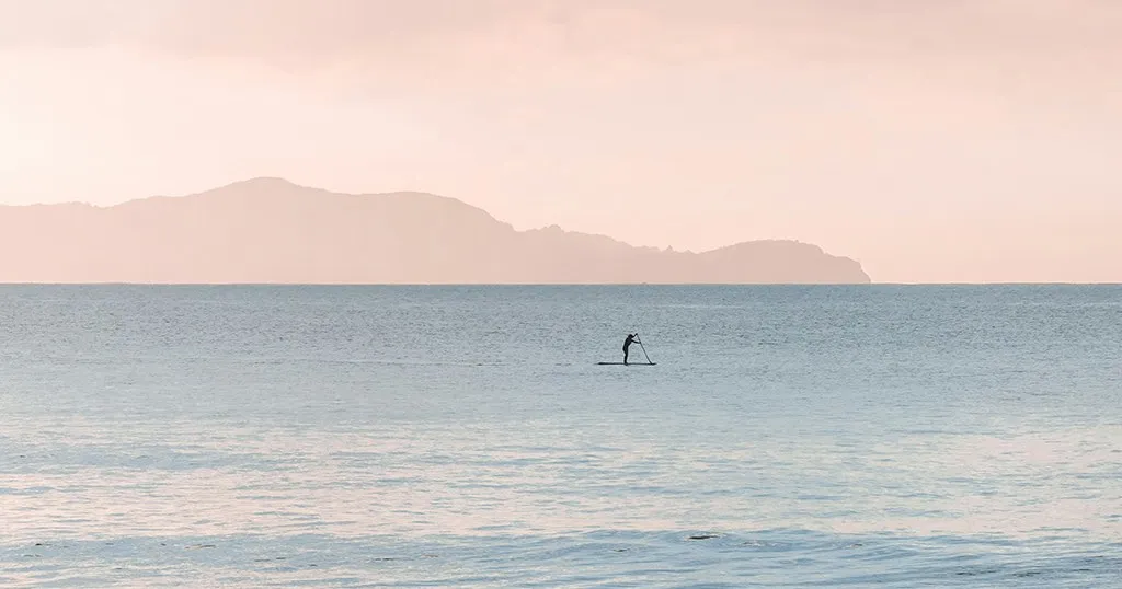 A paddleboarder alone on calm water at dawn, a headland in the pink haze behind — the kind of photograph a good frame is for